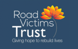 logo for the road victim trust, white writing on a blue background with a flower next to it that has yellow, red and orange petals